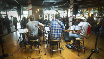 Four musicians performing in a cozy indoor venue. They are seated on stools, playing guitars with music stands in front of them, strumming soulful tunes. The audience in the background is watching attentively. The bar area is visible to the right with warm lighting and holiday decorations.