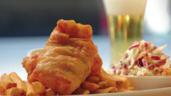 A plate of golden, crispy battered fish served on a bed of thick-cut French fries at Sam's Anchor Cafe. To the side, there is a portion of creamy coleslaw with shredded cabbage and carrots. A tall glass of beer with a foamy head is in the background.