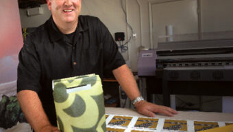 A smiling man in a black shirt stands next to a table displaying printed materials and a decorative, biodegradable bag. Behind him, there is a large printer and various equipment in an eco-friendly workshop or studio setting, emphasizing sustainability.