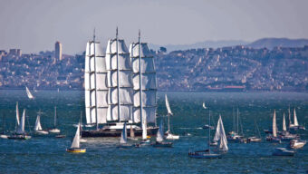 A legendary large black sailboat with white sails is surrounded by numerous smaller sailboats on a body of water. In the background, there is a cityscape with tall buildings and hills. The weather appears clear with calm water, and the sky is light blue.