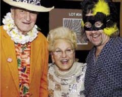Three people dressed in colorful costumes at a party look like they stepped out of the whimsical di Rosa Preserve. The person on the left sports an orange jacket, floral lei, and white hat; the one in the middle dazzles in a sparkly white dress; and the person on the right wears a black and yellow mask.