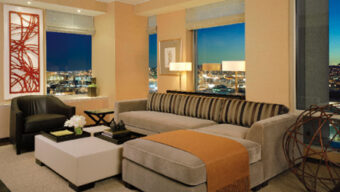 A modern living room with a panoramic city view at night. It features a beige sectional sofa with striped cushions, a brown leather armchair, and a square white ottoman. After its recent makeover, the room now boasts vibrant contemporary decor, including abstract red artwork and geometric sculptures.