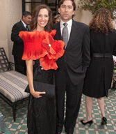 A woman in a vibrant red floral dress holds a black clutch and poses next to a man in a black suit and tie at the Junior League of SF's 83rd Annual Fashion Show. They are standing on a patterned green and white tile floor, while another woman in black is visible in the background.
