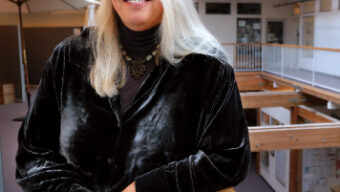 A woman with long, straight, light-colored hair is leaning on a wooden railing in an indoor setting with large windows and exposed wooden beams. Wearing a black velvet jacket and a necklace, she is smiling at the camera, perhaps finding solace after seeking support from Marin Abused Women's Services.