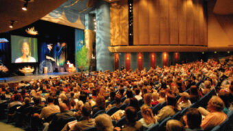 A large audience sits in an auditorium, facing a stage with a speaker. The speaker is projected on a screen for better visibility. The auditorium has dim lighting, high ceilings, and curved rows of seats. The event seems formal, reminiscent of the Bioneers 2007 conference, as attendees are attentively watching.
