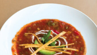 A white bowl of Spain's Soup is garnished with avocado slices, tortilla strips, a drizzle of cream, and a sprig of parsley, on a beige background.