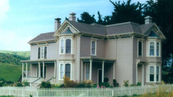 Victorian-style house with beige siding, white trim, and a grey roof, surrounded by a white picket fence. The home features multiple gables, bay windows, and a small porch. It evokes the charm of Historic Ranch Houses and is set against a backdrop of green hills and trees under a clear sky.