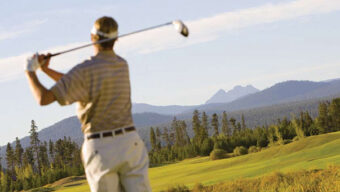 A person swings a golf club on a lush, green golf course with a scenic background of mountains and trees under a clear blue sky. The serene West setting is bathed in natural sunlight as the golfer, wearing a striped shirt and light-colored pants, enjoys the perfect day on prime Golf Real Estate.