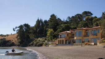 A large, yellow, Mediterranean-style house with arched windows and terracotta roof tiles sits on a beachfront paradise found, surrounded by trees. A person in a small motorboat navigates near the shallow waters of the shore. The sky is clear and blue.