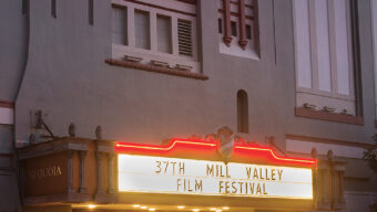 A historic theater with a marquee sign that reads "37th Mill Valley Film Festival" is illuminated at dusk. The building features an ornate facade with art deco elements and large windows. Warm interior light spills out from the entrance, inviting patrons inside to experience Mill Valley's cinematic charm.