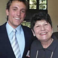 A young man in a suit and tie stands smiling next to an older woman with short, dark hair. The woman, part of a charitable association, is also smiling and dressed in a dark top. They are indoors, and a large window with several panes is visible in the background.