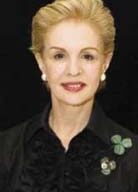 A woman with short, blonde hair and pearl earrings, wearing a black Carolina Herrera top with a ruffled design. She is posing against a black background and has a shamrock-shaped pin on her left side.