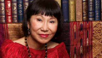 A woman with short black hair and red lipstick is wearing a bright red top and beaded necklace. She is smiling softly, sitting in front of a bookshelf filled with ornate, antique books, among which sits a novel by Amy Tan. The background also features richly patterned textiles.