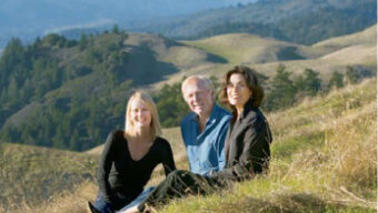 Three people are sitting on a grassy hillside with rolling hills in the background. With Mt. Tam providing a stunning view, the two women are on each side of the man, and all three are smiling at the camera. The scene has an overall sense of tranquility, complemented by the natural landscape behind them.