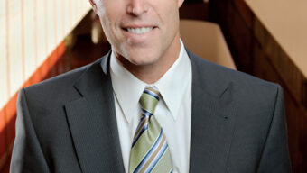 A middle-aged man in a suit and striped tie is smiling at the camera. He has short, light brown hair and bears a resemblance to Jared Huffman. He is standing in a modern building with a long corridor in the background.