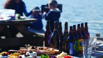 A picnic table by the water is set with various foods and drinks, including salads, cheese, chips, and several beer bottles. In the background, two people, including a child, are seated and appear to be enjoying their time outdoors. It's the perfect setting for memorable picnics.