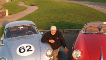A person wearing a cap, known as the resident car guy, kneels between two classic cars parked on a road by a grass field. One car is silver with a large number "62" on the hood, and the other is red. In the background, there is a pathway, more greenery, and some buildings.