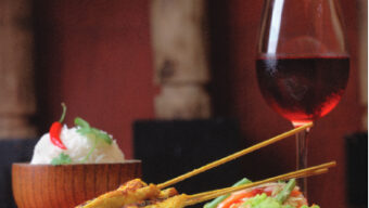 A plated meal from E&O Trading Company consists of skewered grilled meat, a side of salad, and a bowl of white rice garnished with a chili pepper and herbs. A glass of red wine is positioned to the right of the plate. The background is softly lit with warm, earthy tones.