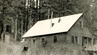 A monochrome image of an old wooden cabin with a steep, snow-covered roof set in a forest. The cabin has a small porch and several windows, reminiscent of the rustic charm found at Marin Ski Club. A tall chimney extends through the roof. Dense trees surround the structure, and an outhouse is visible nearby.