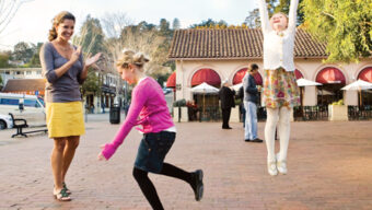 Three people enjoy a sunny day while playing hopscotch on a brick pavement. Spring fever seems to be in the air as a woman claps while a girl in a pink top jumps between squares, and another girl in a white sweater and floral skirt leaps cheerfully with arms raised. A building with red awnings is in the background.