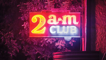 A neon sign with vibrant red and yellow text reading "2AM" and bold blue letters spelling "CLUB" casts a colorful glow against a backdrop of trees and a building wall. The illuminated scene captures the essence of nightlife in a mesmerizing display.