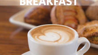 Close-up of a white ceramic cup filled with a latte that has heart-shaped latte art on top, placed on a wooden table. In the background, there is a plate with various pastries. The word "BREAKFAST" is written in bold white letters across the top of this inviting morning scene.