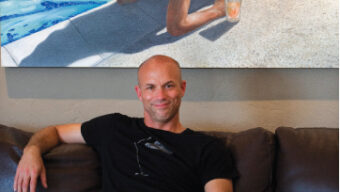A man sits on a brown leather couch in Mill Valley, wearing a black T-shirt and blue jeans. Above him, an Eric Zener painting depicts a woman in a red swimsuit lying by a pool with a drink in hand. The man is smiling with one arm resting on the back of the couch and the other on his lap.