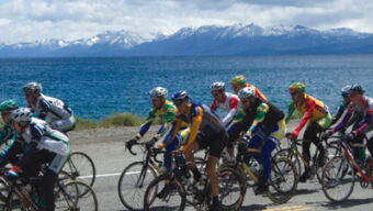 A group of cyclists ride along a road beside a large lake with snow-capped mountains in the background, embarking on their 72-mile journey. They are wearing colorful cycling gear and helmets under a partly cloudy sky.