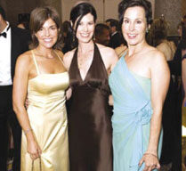 Three women at a formal event, standing close together and smiling. The woman on the left is wearing a yellow gown, the woman in the center has a brown halter dress, and the woman on the right is dressed in a light blue one-shoulder gown. People in the background are celebrating advancements by the Juvenile Diabetes Foundation.