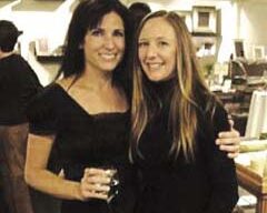 Two women, closely standing together at what seems to be an indoor social event, smile warmly at the camera. The woman on the left, possibly named Taloolah, is holding a drink. Both have long hair and are wearing dark-colored tops. The background reveals other people and various objects.