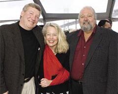 Three people are posing together and smiling. The group includes two men, one on the left and one on the right, and a woman in the center. They are dressed in semi-formal attire, with the woman wearing a red scarf. The background appears to be indoors with large windows, possibly at Epic Roasthouse or Waterbar.