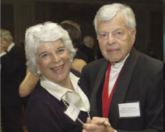 An elderly Dominican couple, dressed formally, smiles and dances together. The woman has white hair and wears a pearl necklace and a name tag, while the man wears a black tuxedo with a red ribbon and a name tag. As honorees, they revel in the warmly lit event setting.