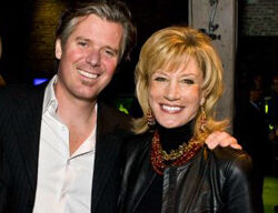 A smiling man and woman pose together in a dimly lit indoor setting at a Buck Institute fundraiser. The man is wearing a dark blazer over a white shirt, while the woman is dressed in a black leather jacket, a black top, and a statement necklace. They appear happy and are standing close to each other.