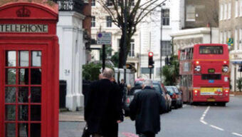 Two elderly men in coats walk on a city sidewalk flanked by a red telephone box. In the distance, a red double-decker bus is visible on the street. Leafless trees and historic buildings line the road, creating a quintessentially British urban scene perfect for London tourism.