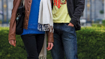 A man and woman, Hansuke Yamamoto and Alexandra McCullagh, stand next to each other smiling outdoors. Alexandra wears a brown jacket, blue top, white scarf, black pants, and black heels. Hansuke sports a yellow graphic t-shirt, dark jacket, blue jeans, and gray sneakers. A building and greenery are in the background.