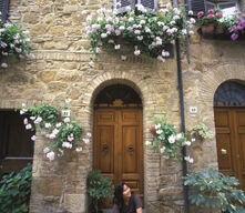 A person sits on a doorstep in front of a stone building adorned with green plants and blooming pink and white flowers. With wooden doors and shuttered windows adding to the charming, rustic ambiance, the scene feels like a Tuscan infusion of timeless elegance and natural beauty.