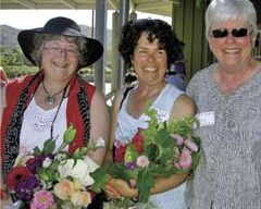 Three smiling women stand outdoors, each holding a colorful bouquet of flowers. The woman on the left wears a wide-brimmed hat and red scarf, the middle woman with curly hair is dressed in white, and the woman on the right, wearing sunglasses and a grey top, proudly represents Marin Horizon School.
