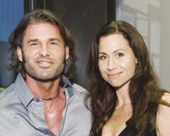 A man and woman are posing together, both smiling. The man has shoulder-length dark hair, wears an open-buttoned shirt, and has a necklace. The woman has long dark hair and is wearing a sleeveless top. They appear to be at Barney's Grand Opening event indoors.