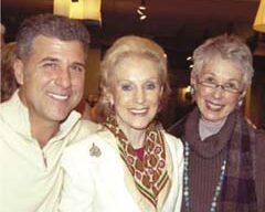A man and two women are smiling at the camera in a warmly lit, NapaStyle indoor setting. The man is on the left, in a white shirt. The woman in the middle sports a white jacket and patterned scarf. The woman on the right wears glasses and a dark outfit complemented by a beaded necklace.