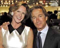 A man in a suit with a blue tie stands next to a woman in a sleeveless white dress. He has his arm around her shoulder as they both smile, with string lights in the background hinting at a festive event, perhaps an antiques show in San Francisco.