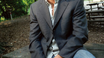 A man in a gray blazer and blue jeans, resembling Helge Hellberg, sits on a picnic table in a wooded park. He has short, dark hair and is looking into the camera with a slight smile. Trees and another picnic table are visible in the background.
