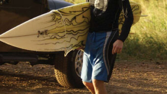 A person wearing a black rash guard and blue board shorts, emblazoned with "Fear the Shark," is carrying a white surfboard with a wavy design. They are walking barefoot on a dirt path next to a parked car, with greenery and other vehicles visible in the background.