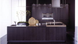 Modern kitchen with dark cabinets and flooring. Features a central island with a sink, stovetop, and decorative baskets. Stainless steel appliances are mounted in the back wall, with a minimalist, sleek design. Abundant natural light illuminates the room from the left side, highlighting the beautiful kitchen islands.