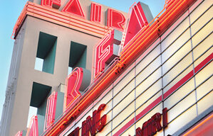 An old-fashioned movie theater marquee with red neon lights displays the names of two films, "Changeling" and "Zack & Miri." The marquee, underlined by the theater name "Fairfax" illuminated in red neon lights, stands out against the clear blue sky.