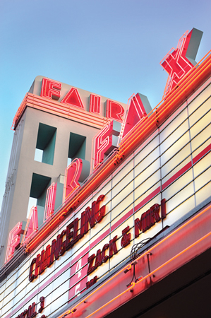 An old-fashioned movie theater marquee with red neon lights displays the names of two films, "Changeling" and "Zack & Miri." The marquee, underlined by the theater name "Fairfax" illuminated in red neon lights, stands out against the clear blue sky.