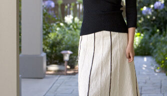 A woman with long, wavy hair smiles while standing next to a column on a tiled walkway. She is wearing an exquisite Anyi Lu black off-the-shoulder top, paired with a knee-length white skirt adorned with dark vertical lines, and elegant black heels. The background features greenery and out-of-focus flowers.