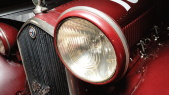 Close-up shot of a vintage red car's front end, featuring a large round headlight, chrome detailing, and the brand emblem above the grille. The bodywork is sleek, with visible latches and vents, reflecting the classic design of cars that still sparks automotive talk today.