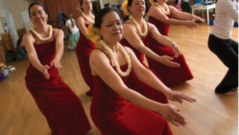 A group of people wearing red dresses and yellow flower leis perform a lovely hula dance in a room with wooden floors and a large mirror on the wall. They extend their hands forward while bent at the knees, focusing intently on their movements.