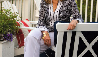 A woman with blonde hair, wearing a white and black patterned top and white pants, sits on the armrest of a bench on a deck. The deck, which resembles a Belvedere with its railing and potted plant with purple flowers, overlooks trees. She is smiling at the camera.