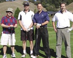 Four men are standing on a golf course with golf clubs, posing for a picture after a friendly tournament. They are dressed in casual golf attire, including polo shirts, shorts, and golf shoes. The background features rolling hills, trees, and clear skies.
